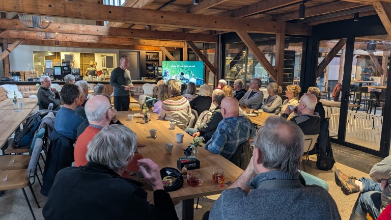 Groep mensen luistert naar een presentatie in een houten zaal met een scherm waarop een diapresentatie te zien is.