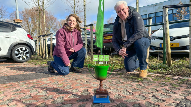 Twee mensen gehurkt naast een groene trofee met pictogrammen, op een parkeerplaats met auto's en bomen op de achtergrond.