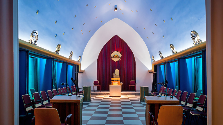 Vergaderzaal met geblokte vloer, centrale tafel, rode stoelen, en een oogmotief op een gordijn onder een blauwe gewelfde plafond.