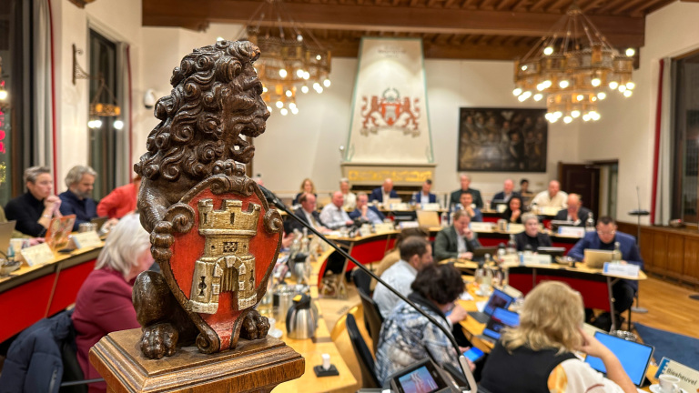 Een vergaderruimte met een beeld van een leeuw met een schild op de voorgrond, en meerdere mensen zittend aan tafels op de achtergrond, verlicht door kroonluchters.