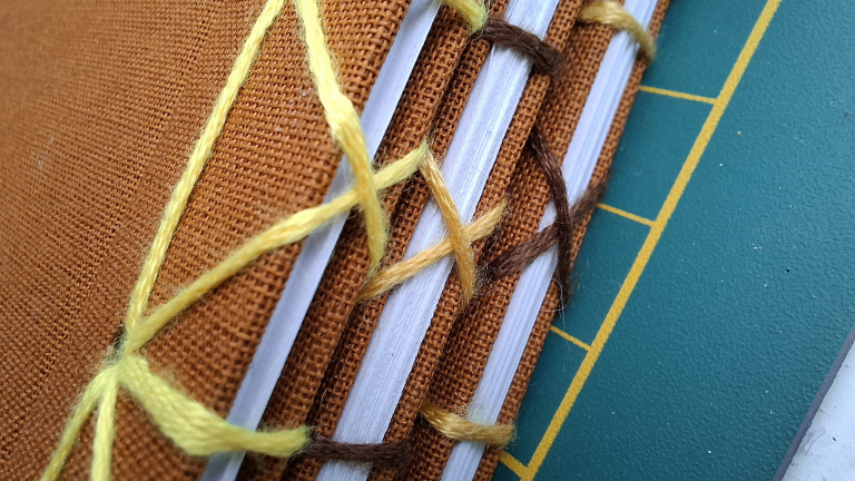 Close-up van een boek met oranje kaft, genaaid met bruine en gele draad, op een groene ondergrond met gele lijnen.