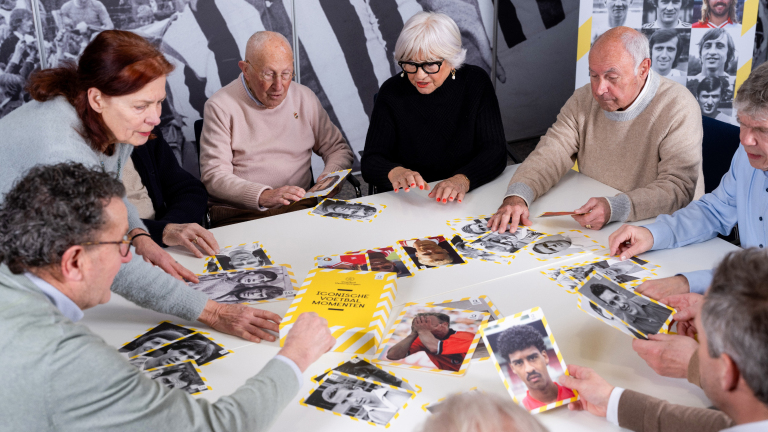 Groep oudere mensen zit rond een tafel met foto's van historische voetballers voor zich uitgestald.