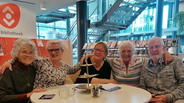 Vijf lachende ouderen zitten aan een tafel in een bibliotheek met een tijdschriftenrek op de achtergrond.