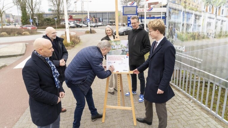 Mensen staan rondom een bord dat wordt ondertekend op een stadsplein, met een achtergrond van winkels en een informatiebord.