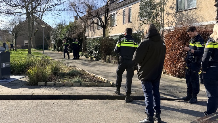 Politieagenten in uniform staan bij een woonwijk en overleggen, met een afvalcontainer en een rij huizen op de achtergrond.