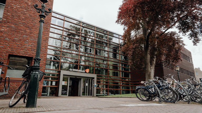 Vooraanzicht van een moderne bibliotheek met glazen gevels, omgeven door fietsen en een grote boom.