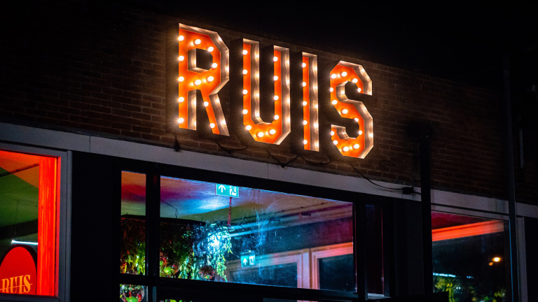 Verlicht bord met het woord "RUIS" boven een gebouw met ramen en kleurrijke verlichting binnenin.