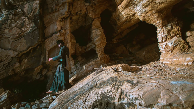 Een persoon loopt langs een rotswand met grotten, gekleed in donkere kleding en een tas dragend.