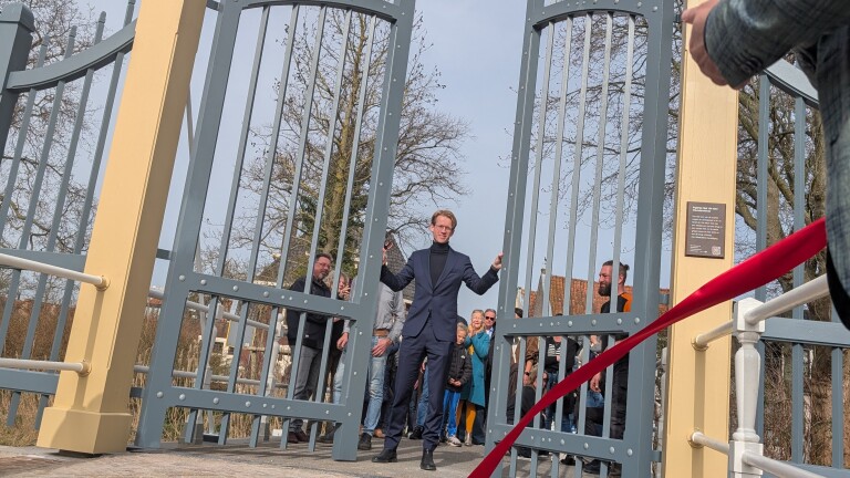 Een man in een blauw pak opent een groot smeedijzeren hek, omringd door toeschouwers, met een rode lint zichtbaar op de voorgrond.