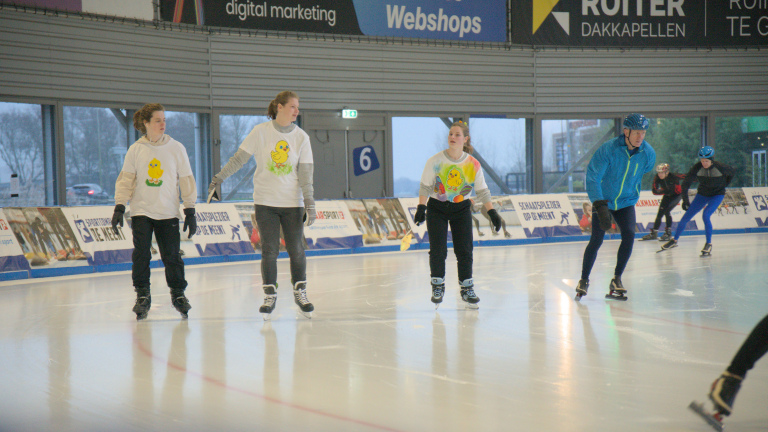 Mensen schaatsen in een indoor schaatsbaan, sommigen dragen shirts met een gele eend.