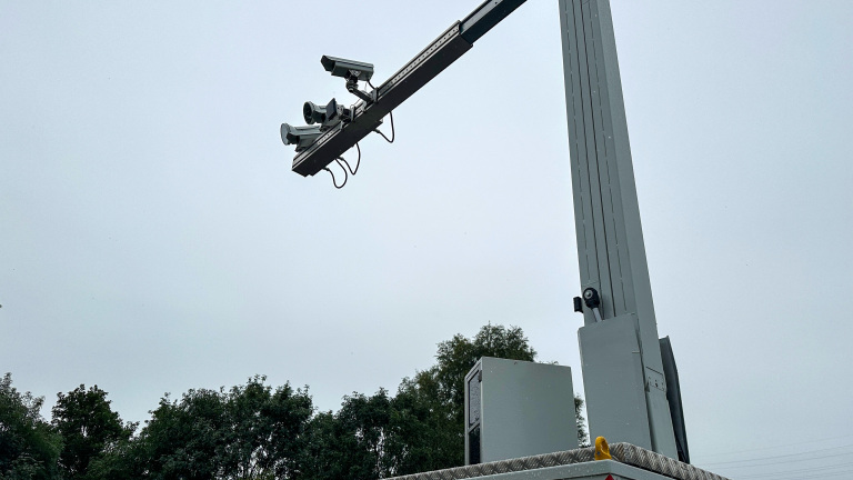 Verkeerscamera's gemonteerd op een hoge mast tegen een bewolkte hemel, met bomen op de achtergrond.