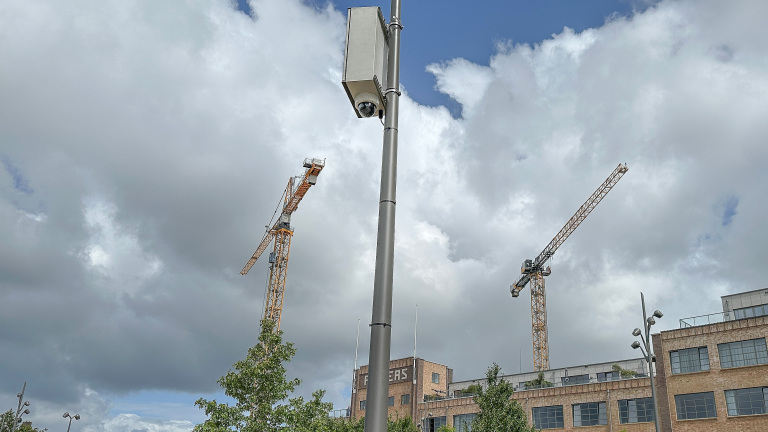Een beveiligingscamera hangt aan een hoge paal met op de achtergrond twee grote bouwkranen en een bakstenen gebouw, onder een bewolkte lucht.