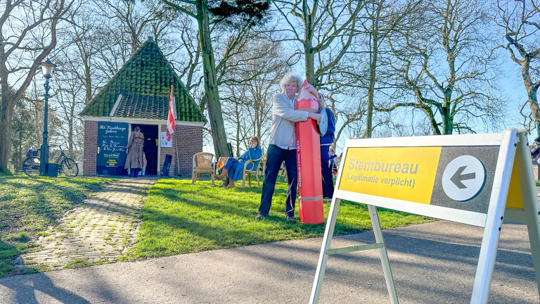 Man omarmt een groot rood potlood naast een bord met "Stembureau (Legitimatie verplicht)" voor een klein huisje.