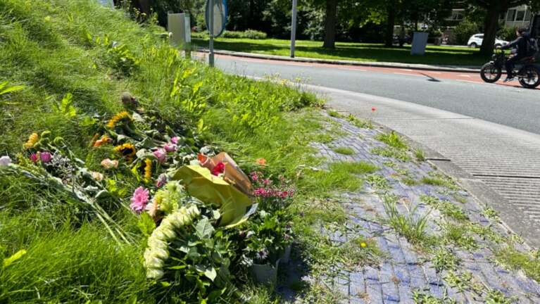 Bloemen leggend langs een weg, mogelijk een herdenkingsplek, met op de achtergrond een fietser op een zonnige dag.