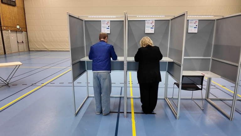 Twee mensen stemmen in stemhokjes in een gymzaal.