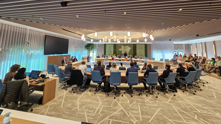 Grote vergaderruimte met mensen zittend aan een ronde tafel, moderne inrichting en scherm aan de muur.