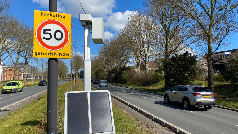 Een verkeersbord met een snelheidslimiet van 50 km/u, gemarkeerd als herhaling en geluidshinder, naast een drukke weg met auto's en een geluidsmeetpaal, omgeven door bomen en enkele huizen.