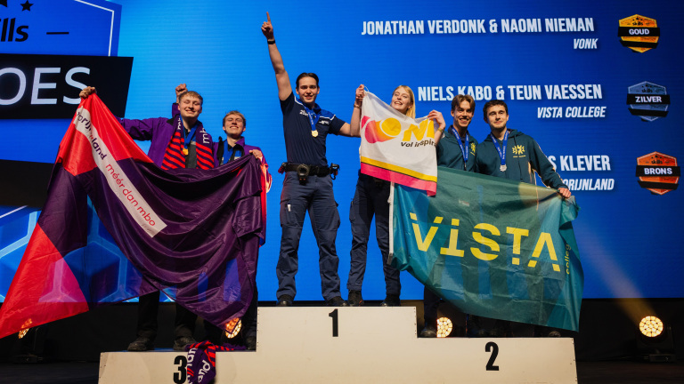 Winnaars op het podium met medailles en vlaggen, omringd door een blauw scherm met namen en awards in goud, zilver, en brons.
