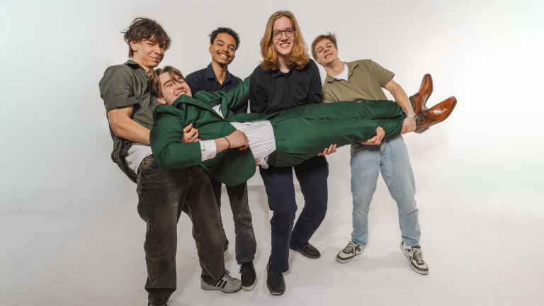 Vier jonge mannen tillen lachend een andere man in een groen pak op in een studio.