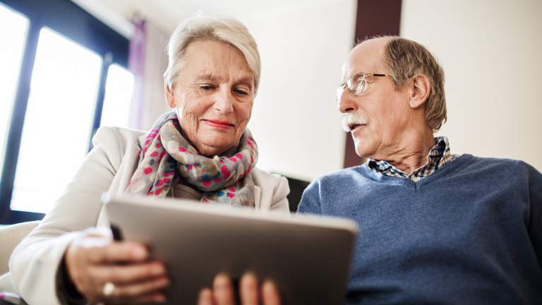 Ouder koppel zit op een bank en gebruikt een tablet, de vrouw draagt een sjaal met stippen en de man een bril.