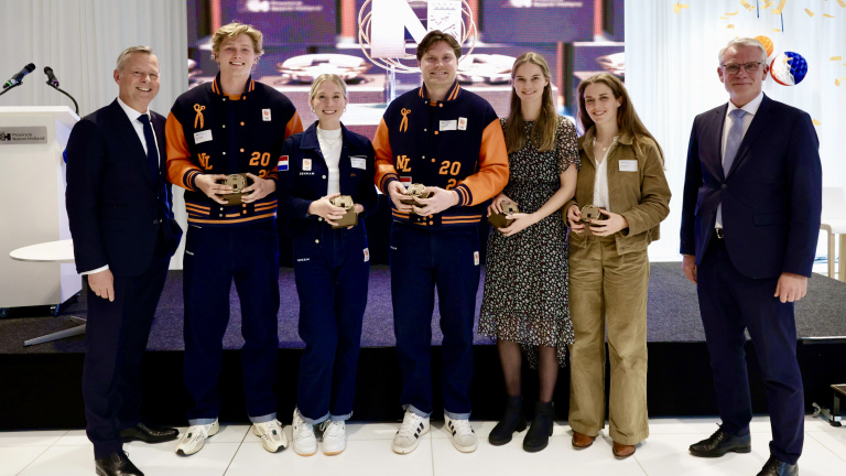 Zes mensen poseren samen, drie dragen sportjassen en houden trofeeën vast, naast twee nette mannen.
