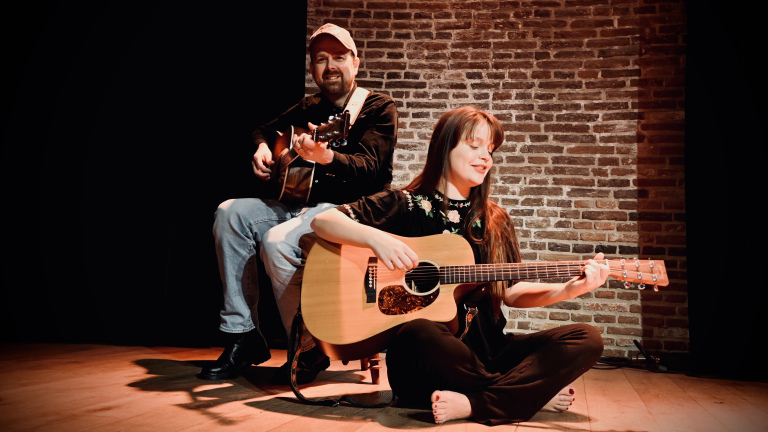 Twee muzikanten spelen gitaar op een podium met een bakstenen achtergrond; de vrouw zit en de man zit op een kruk.