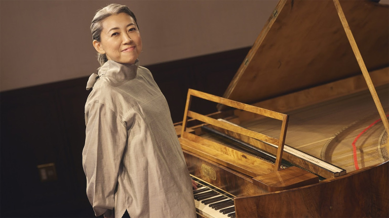 Een persoon met grijs haar glimlacht naast een houten piano met de klep omhoog in een sfeervolle ruimte.