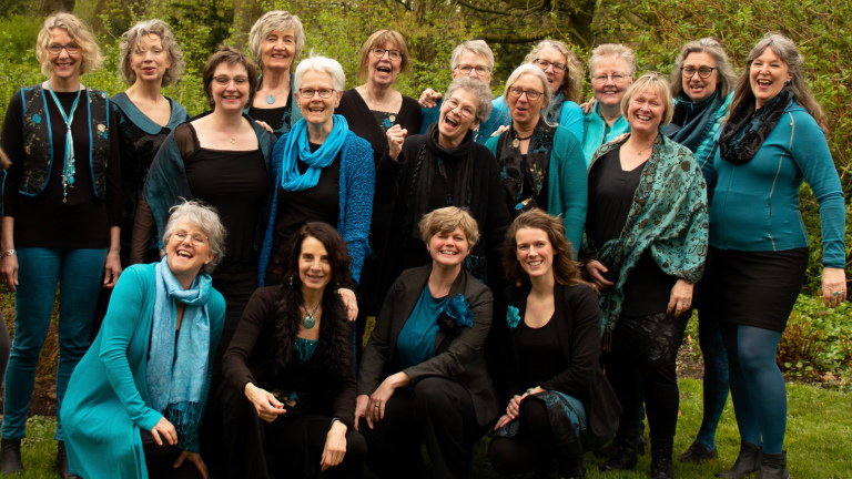 Groep lachende vrouwen in blauwe en zwarte kleding poseren buiten voor een groene achtergrond.
