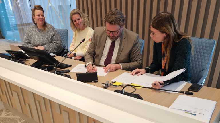 Vier personen zitten aan een tafel; een man in pak ondertekent een document, terwijl de anderen toekijken.