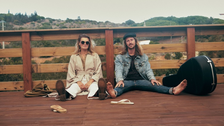 Twee mensen zitten ontspannen op een houten terras, met een gitaarkoffer naast hen en een natuurlijke achtergrond.