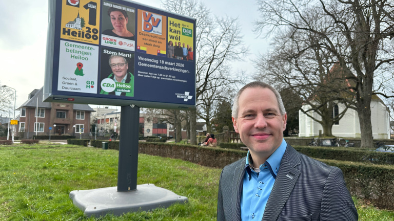 Man poseert voor campagnebord van gemeenteraadsverkiezingen Helmond, met informatie en gezichten van verschillende politieke partijen.