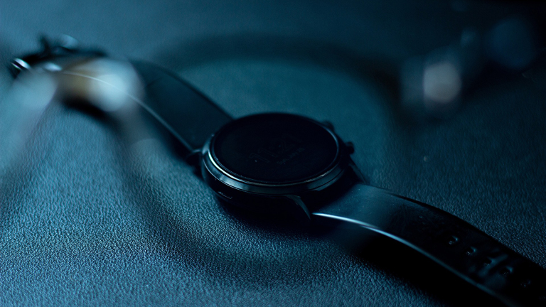 Een zwarte polshorloge met leren band ligt op een donkere ondergrond, deels wazig door een voorgrondobject.