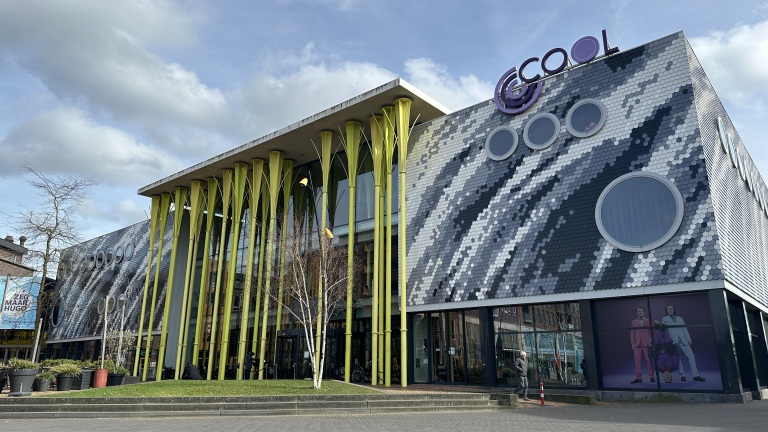 Moderne gebouw met kleurrijke, gele pilaren en een gevel met schubbenpatroon en ronde ramen, onder een blauwe lucht.