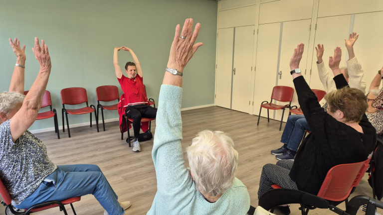 Ouderen doen zitoefeningen in een kring terwijl een begeleider in een rode trui de beweging voordoet.