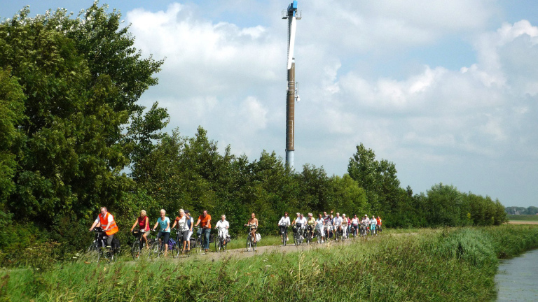 Grote groep fietsers rijdt langs een groen pad met bomen en een hoge toren op de achtergrond.