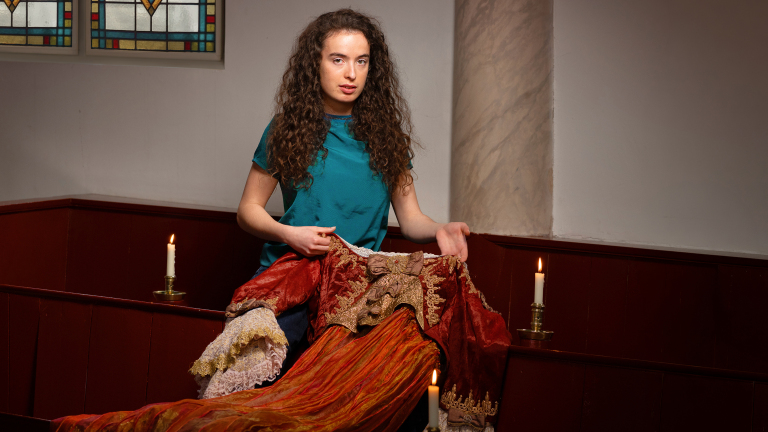 Vrouw met krullend haar houdt een rijkelijk versierde rode jurk vast, zittend in een kamer met brandende kaarsen en glas-in-loodramen.