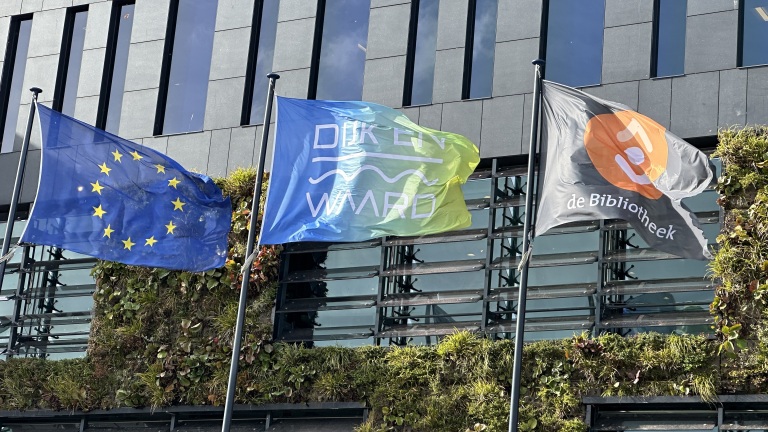 Drie vlaggen wapperen voor een gebouw met verticale tuinen: de EU-vlag, "Dijk en Waard," en de Nederlandse bibliotheekvlag.
