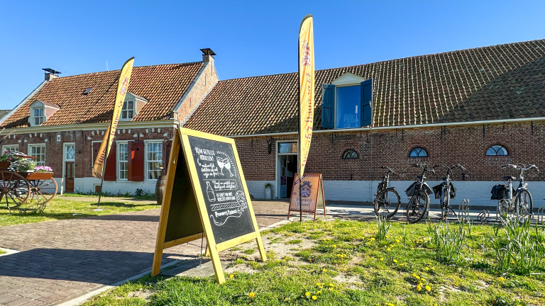 Bakstenen gebouw met schuine dakpannen, fietsenrekken, en een bord met opschrift voor een brouwerij, omringd door zonnig gras.