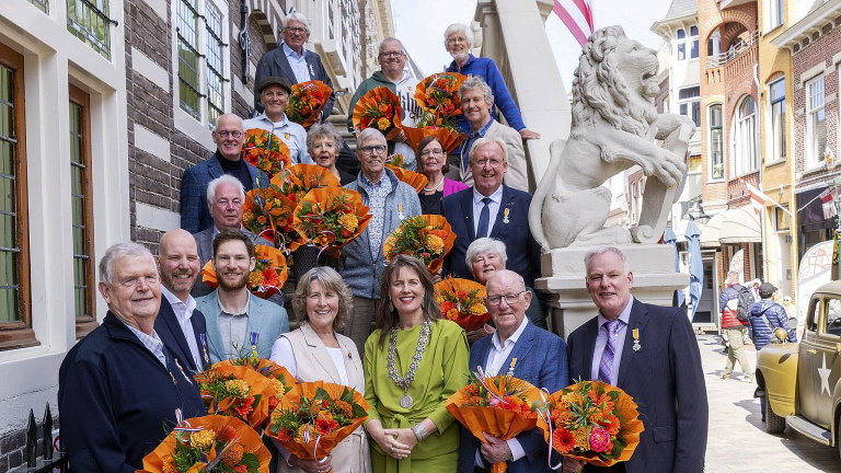Een groep mensen poseert met boeketten voor een gebouw, sommigen dragen medailles.
