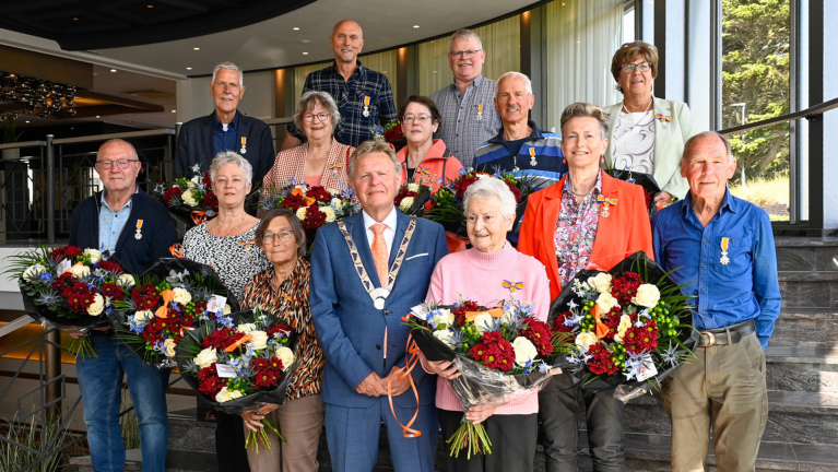 Een groep mensen die lintjes en grote boeketten bloemen vasthouden, staat samen met een persoon in een ambtsketen op een trap binnenshuis.