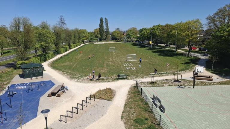 Speelveld met voetbaldoelen en kinderen die spelen, omgeven door bomen en paden, met een basketbalveld en speeltoestellen.