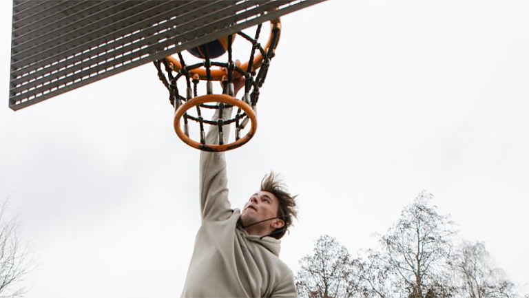 Persoon die springt om een basketbal in een basketbalring te dunken, met bomen op de achtergrond.