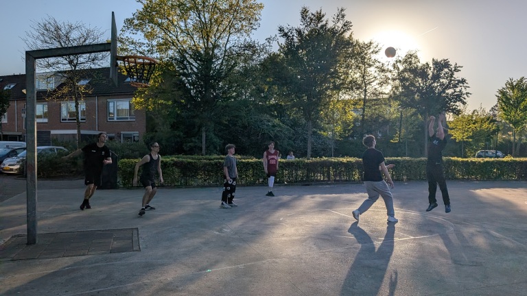 Mensen spelen basketbal op een buitenveld, omgeven door bomen en huizen, met de zon laag aan de hemel.