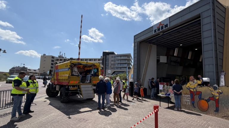 Mensen staan buiten een KNRM-station naast een voertuig met open achterdeur; het gebouw heeft een open garagedeur en een affiche.