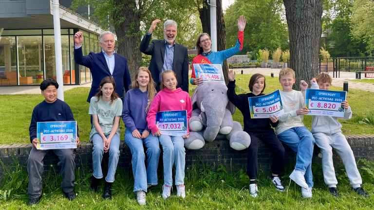 Een groep mensen en kinderen zit buiten op een muurtje, elk met borden die de opbrengst van een inzamelingsactie tonen.