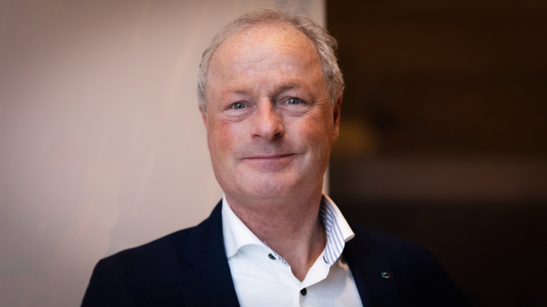 Man met grijs haar en jasje, glimlachend naar de camera in een neutrale omgeving.