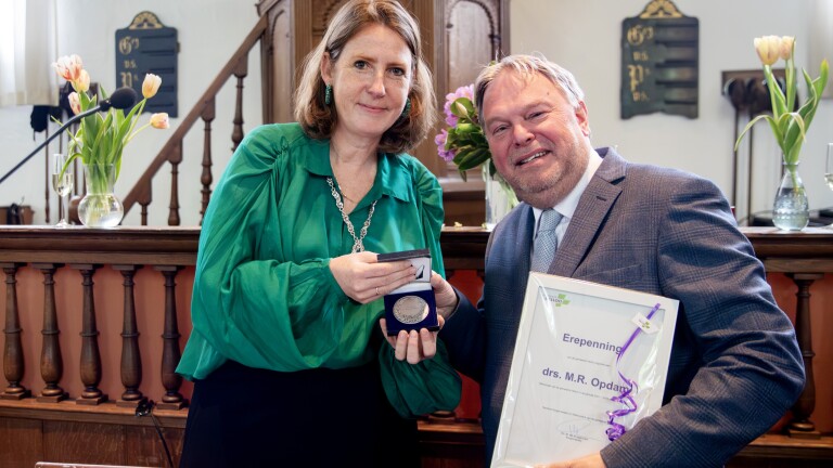 Twee mensen poseren met een medaille en een oorkonde in een ceremonieel decor met een katheder en bloemen.