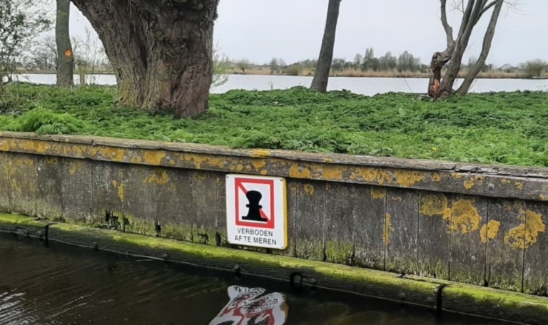Muur langs een waterkant met een bord "Verboden af te meren" en een achtergrond van groene vegetatie en bomen.
