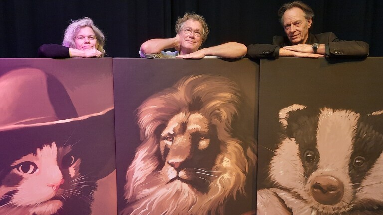 Drie mensen leunen op panelen met schilderijen van een kat, leeuw en das tegen een donkere achtergrond.