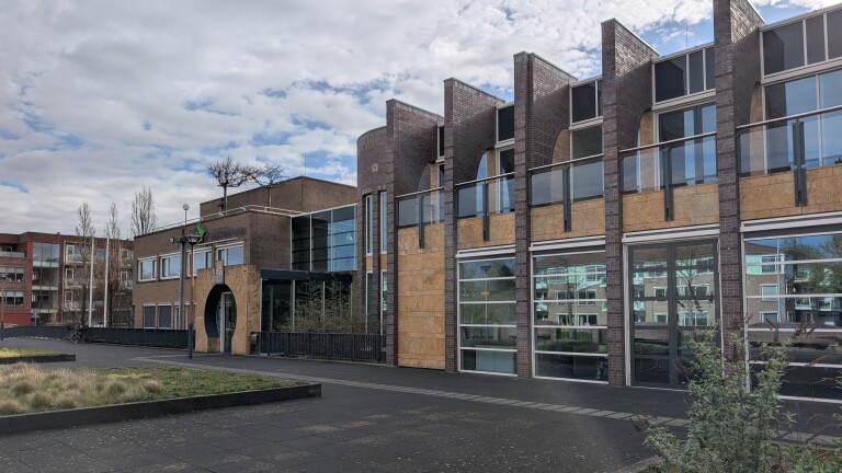 Modern gebouw met grote ramen, bakstenen facade en geometrische vormen onder een bewolkte hemel.